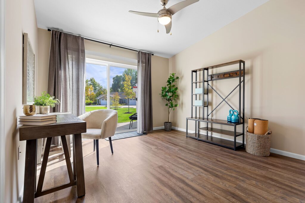 A bright, modern room with wood flooring, a desk and chair by a window, open shelving, a potted plant, and neutral decor. Large glass doors open to a grassy yard.