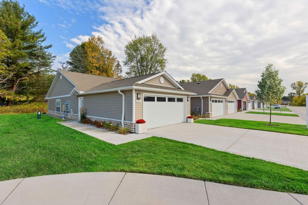 Row of single-story suburban houses with attached garages, gray siding, and well-kept lawns on a sunny day.