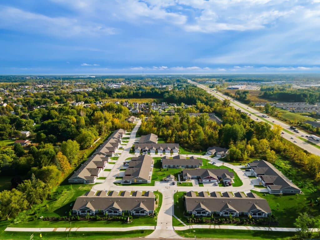 Aerial view of a suburban residential neighborhood with rows of houses, green lawns, trees, and a highway running alongside the area under a partly cloudy sky.