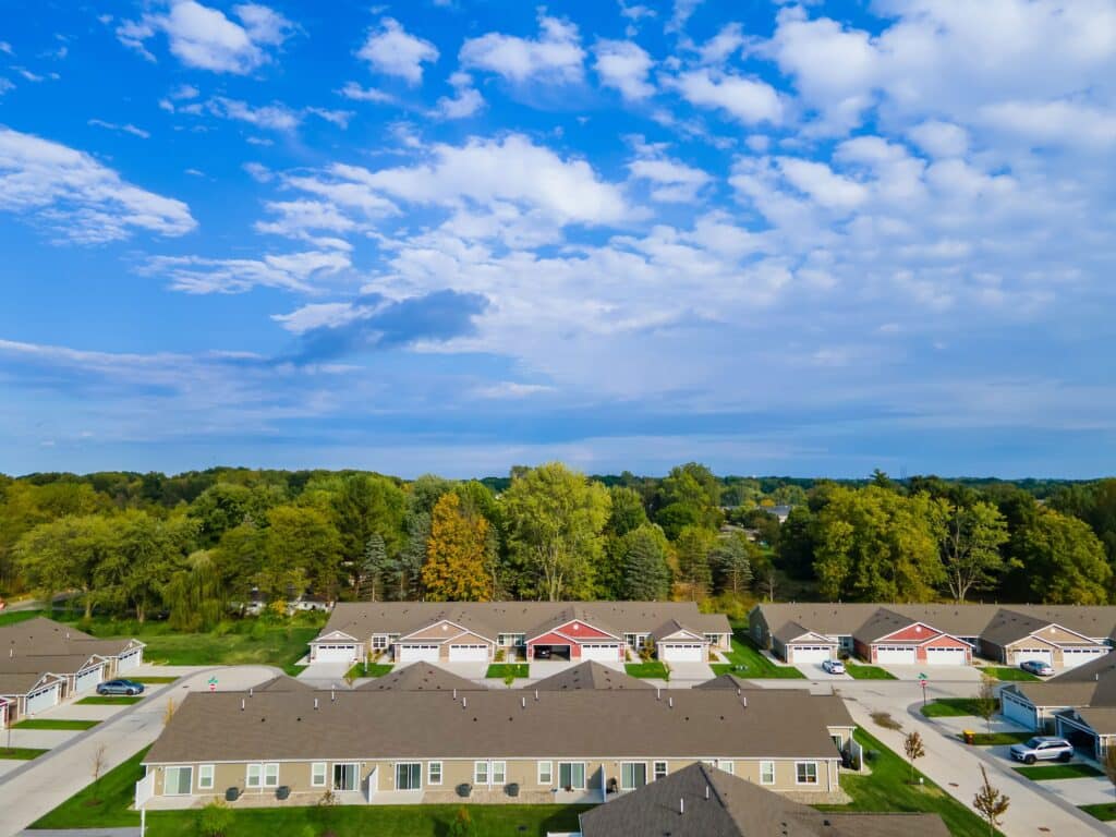 A row of single-story houses with garages is surrounded by trees, with a blue sky and scattered clouds overhead.