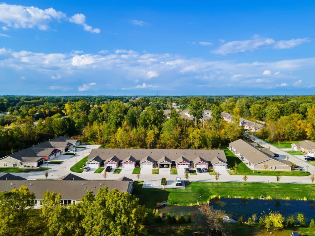 Aerial view of a suburban neighborhood with rows of houses, driveways, trees, and a small pond in the foreground under a blue sky.