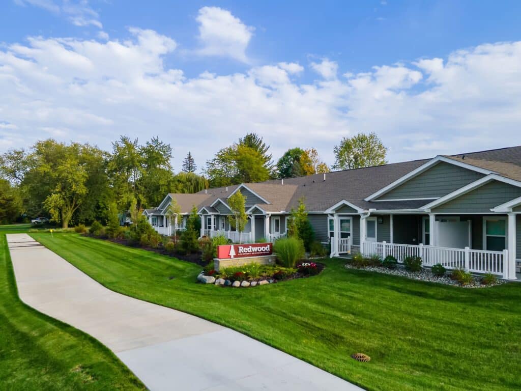 A row of single-story residential units with gray siding and white trim, well-maintained lawns, landscaping, and a sidewalk, with a sign reading "Redwood" in front.