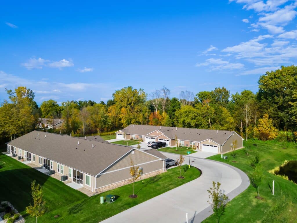 Aerial view of a residential complex with single-story buildings, green lawns, a curved driveway, parked cars, and trees in the background under a clear blue sky.