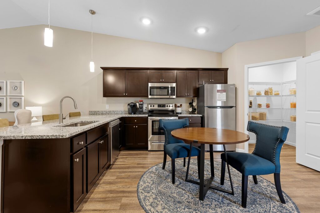 Modern kitchen with dark wood cabinets, stainless steel appliances, a round dining table with two blue chairs, and a walk-in pantry with shelves in the background.