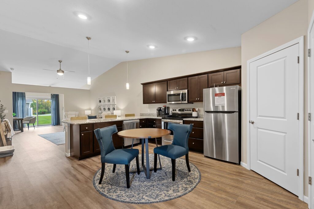 Modern kitchen with dark wood cabinets, stainless steel appliances, a round table with two blue chairs on a rug, and open layout leading to a living area with glass doors.