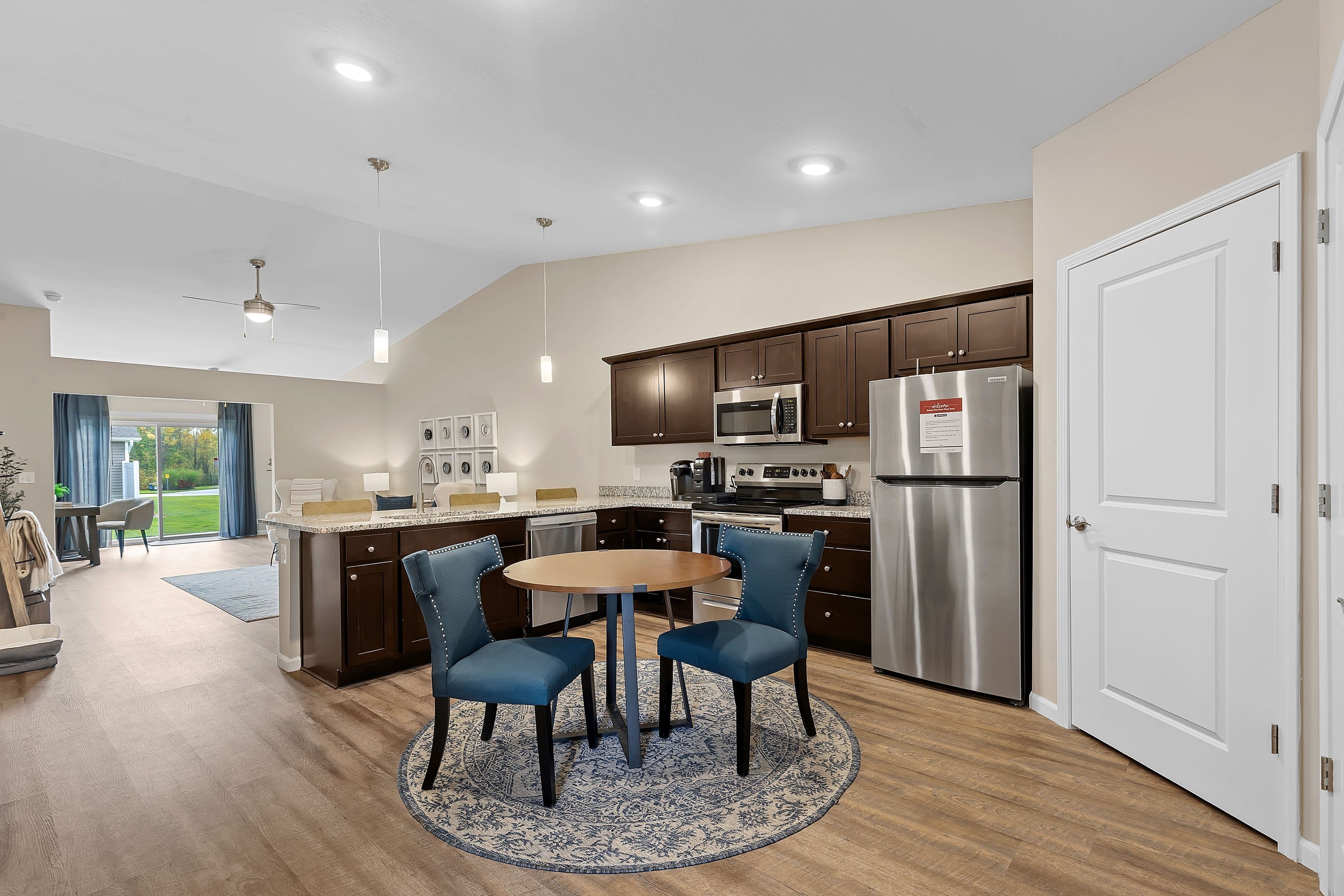Modern kitchen with dark wood cabinets, stainless steel appliances, a round table with two blue chairs on a rug, and open layout leading to a living area with glass doors.