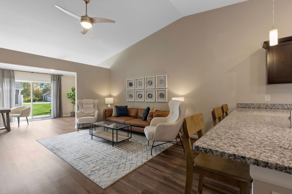 Modern living room with a brown sofa, two armchairs, a coffee table, and wall art, adjacent to a kitchen counter and large windows overlooking a grassy yard.