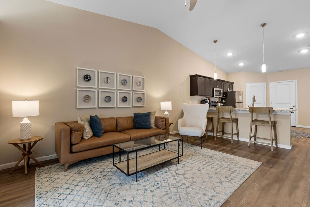 A modern living room with a tan sofa, glass coffee table, armchair, and open kitchen with bar stools under pendant lights on a wood floor.