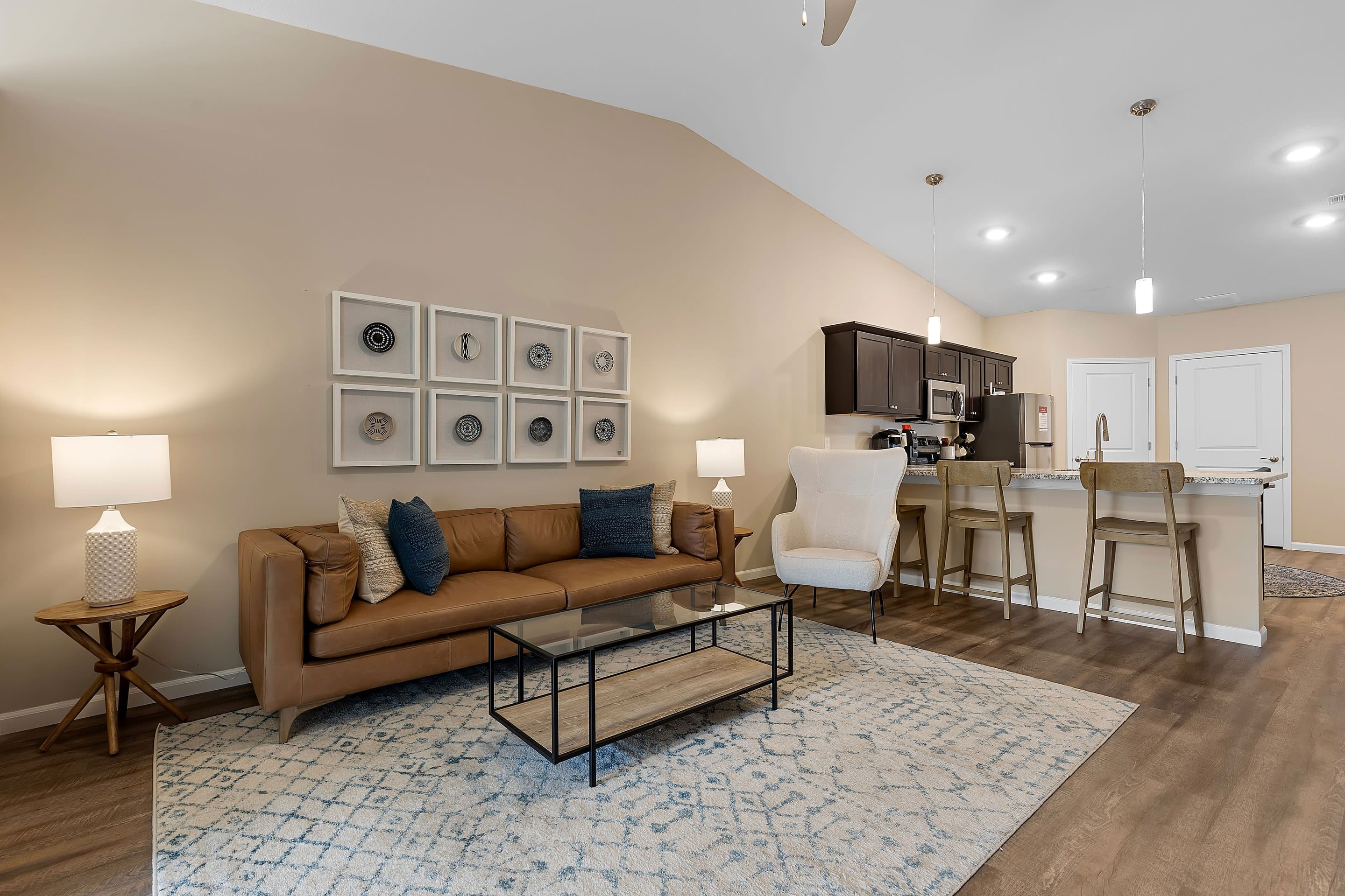 A modern living room with a tan sofa, glass coffee table, armchair, and open kitchen with bar stools under pendant lights on a wood floor.