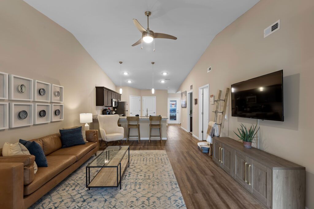 Open-concept living room and kitchen with brown leather sofa, armchair, glass coffee table, wall-mounted TV, kitchen island with barstools, and wood flooring.