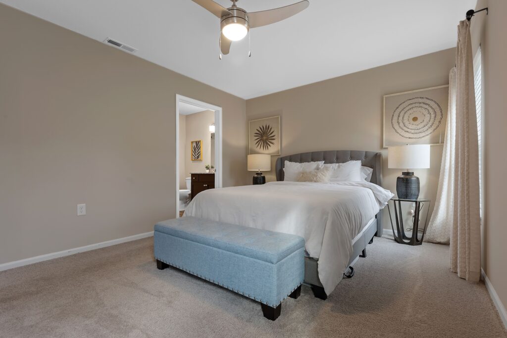 A bedroom with a bed, white bedding, two lamps, framed wall art, a blue upholstered bench, beige carpet, and an open door leading to a bathroom.
