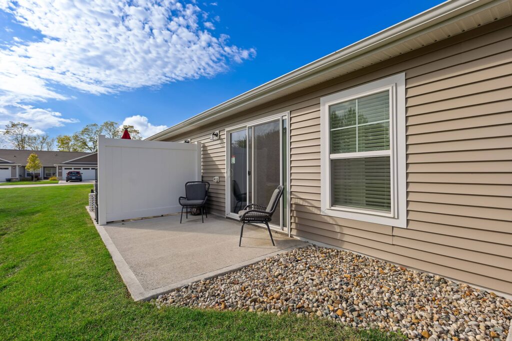 A small concrete patio with two black chairs, a white privacy screen, and tan siding on a single-story house, overlooking a grassy yard and driveway.
