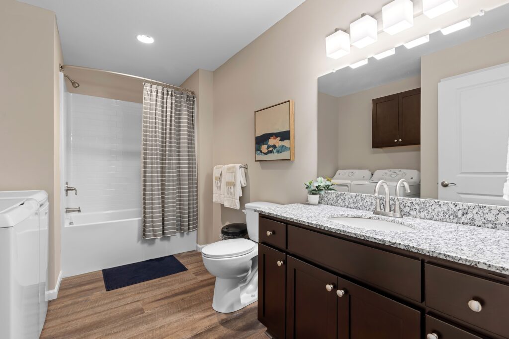 Modern bathroom with granite countertop, dark wood vanity, toilet, bathtub with striped shower curtain, washer and dryer, and wall art above a towel rack.