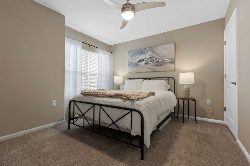 A neatly made bed with white linens, two bedside tables with lamps, a landscape painting above the bed, and a ceiling fan in a beige-walled bedroom with carpeted floor.