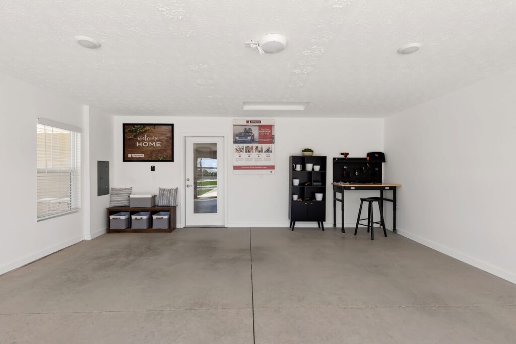 A clean, organized garage with a door and window, wall art, storage bins, a black shelving unit, and a small workbench with two stools against a white wall.
