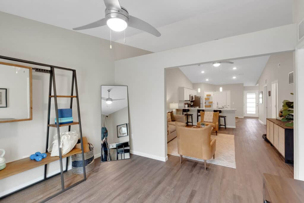 Modern open-concept living space with neutral walls, wood flooring, a ceiling fan, and a view of the kitchen and living room featuring tan chairs and a dining area.