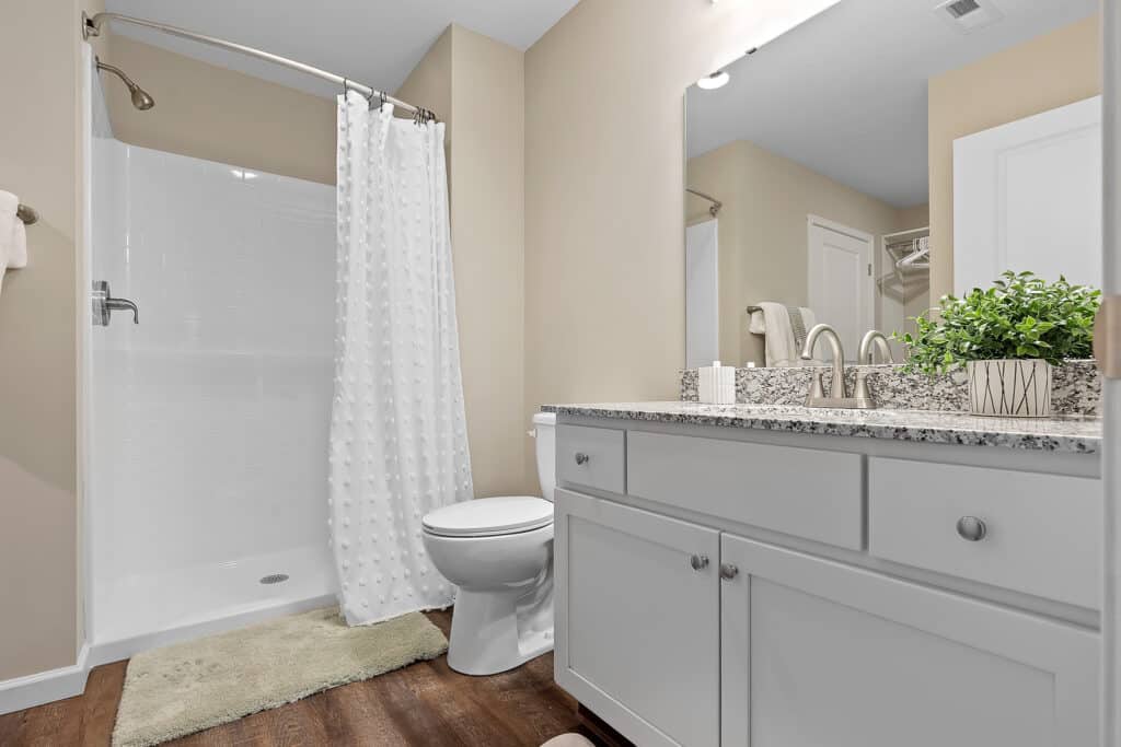 Modern bathroom with a walk-in shower, white shower curtain, toilet, light granite countertop, white vanity, and a potted plant on the sink.