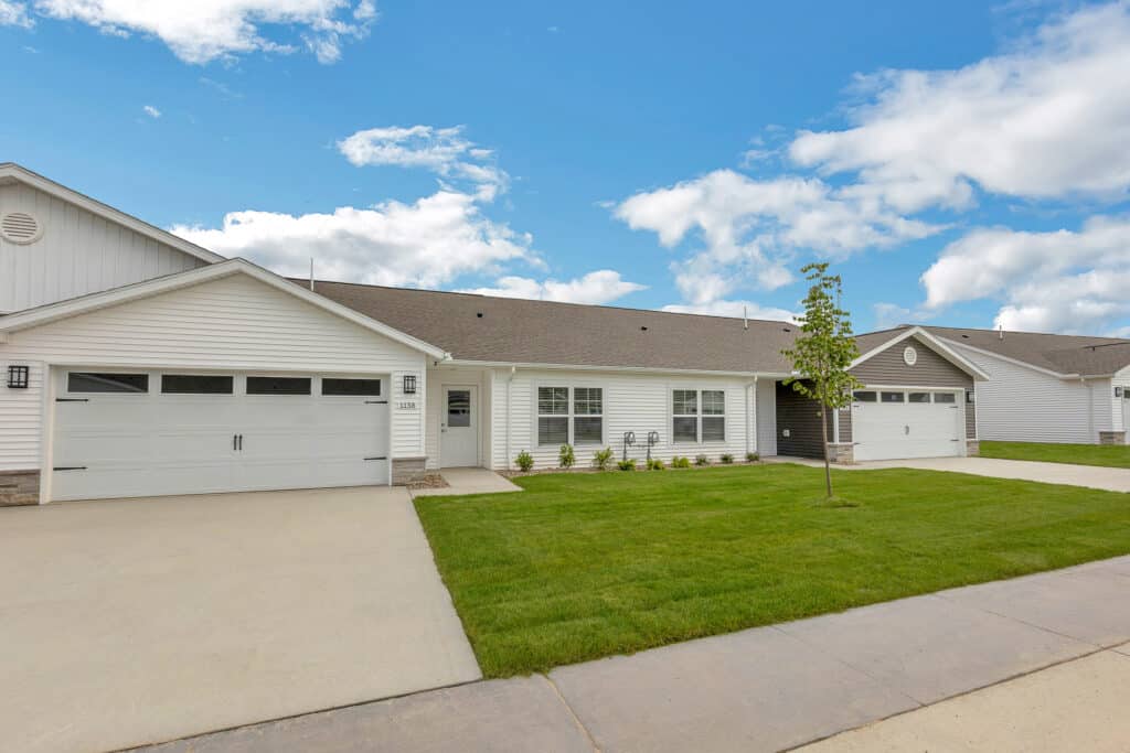 Single-story white house with a double garage, small front lawn, young tree, and a clear blue sky with scattered clouds.