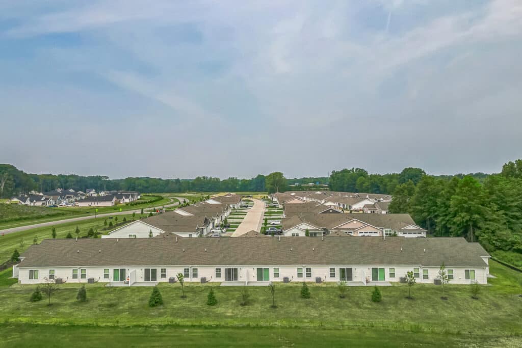 Aerial view of a suburban neighborhood with a row of single-story houses in the foreground, a road to the left, and trees in the background under a cloudy sky.