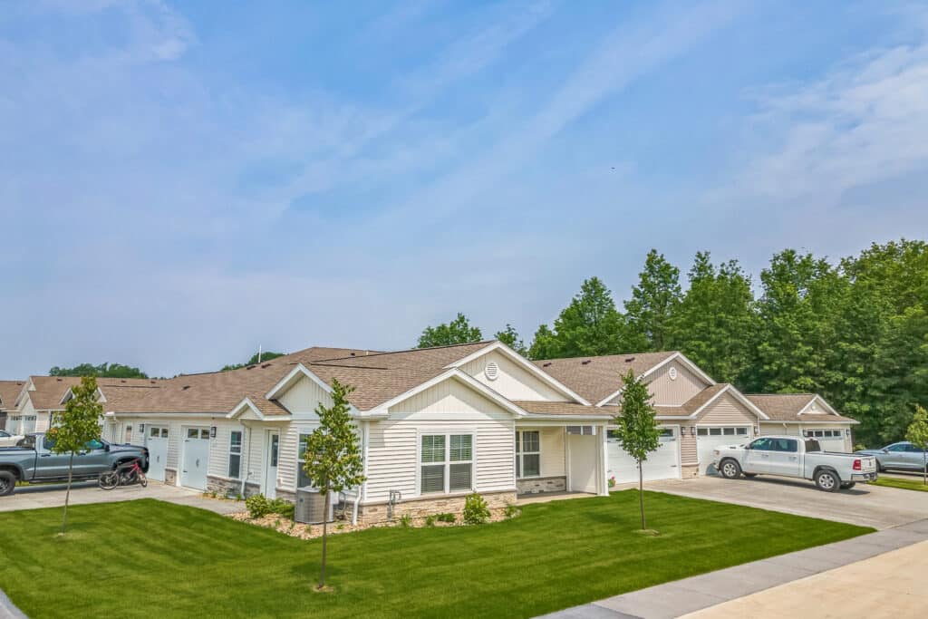 Single-story white duplex homes with attached garages, green lawns, young trees, parked vehicles, and a clear sky in a suburban neighborhood.