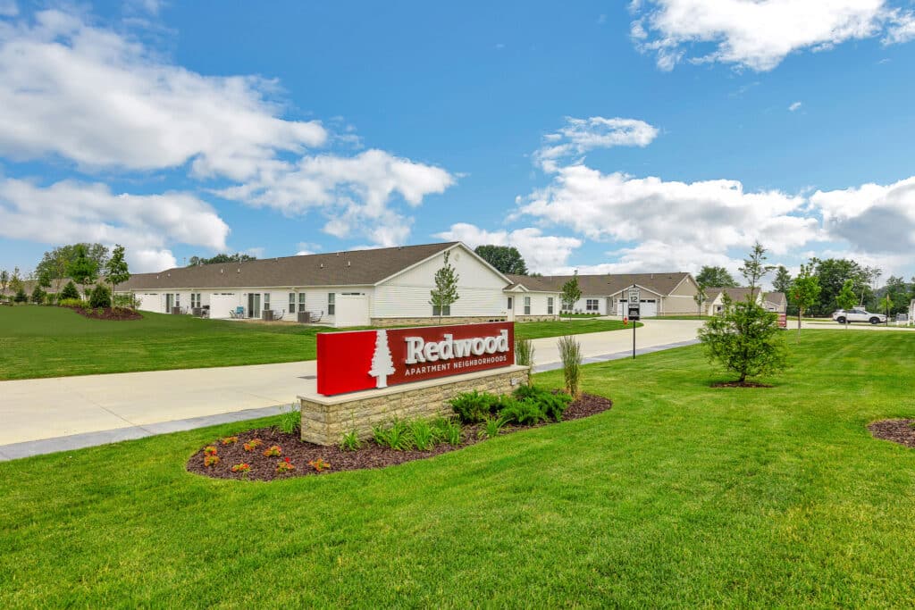 A landscaped lawn features a red and white Redwood Apartment Neighborhood sign in front of single-story residential buildings under a partly cloudy sky.