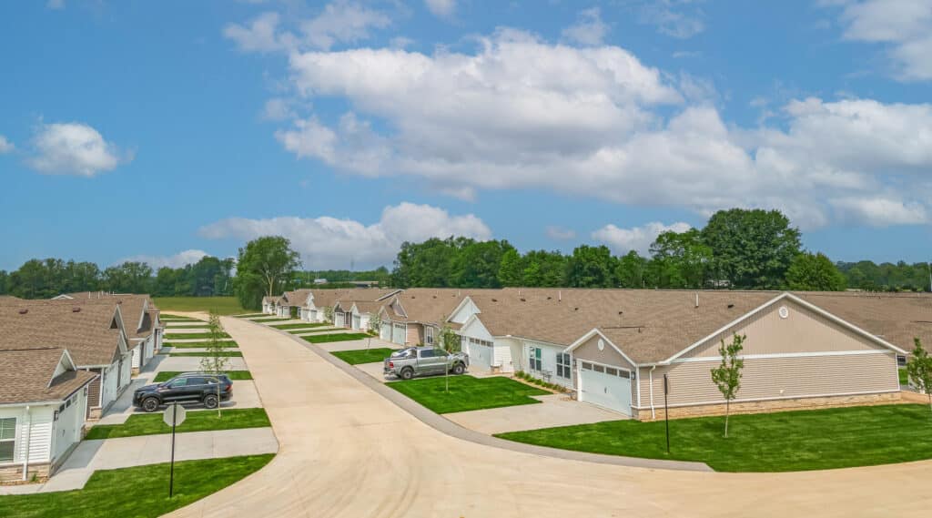 A row of single-story suburban houses with garages, driveways, and parked cars along a clean, curved street under a partly cloudy sky.