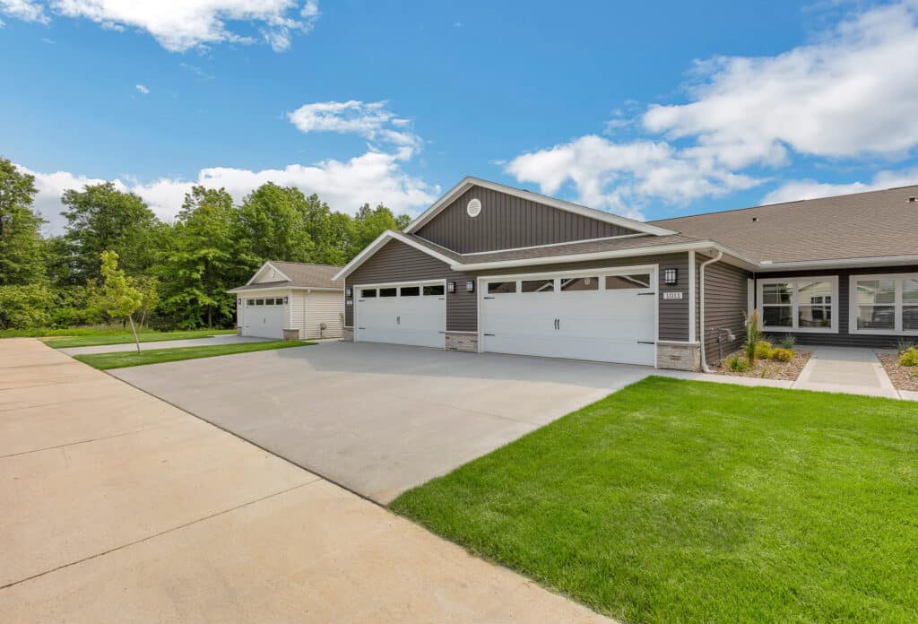 A row of modern single-story homes with gray siding, large driveways, and double garages under a partly cloudy blue sky.