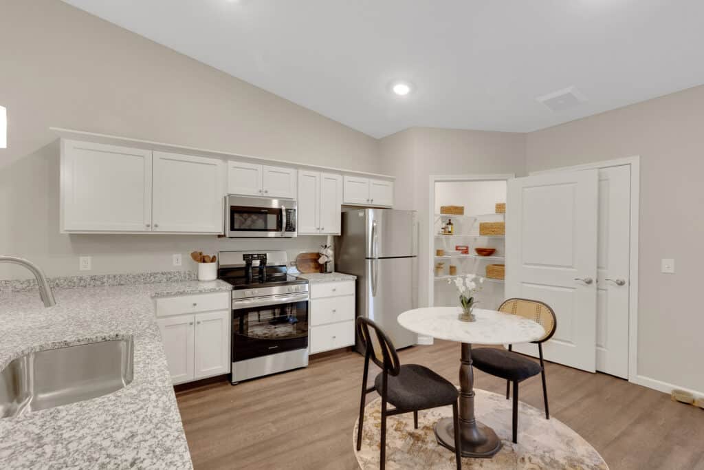 Modern kitchen with white cabinets, stainless steel appliances, a round dining table with two chairs, and an open pantry with shelves containing baskets.