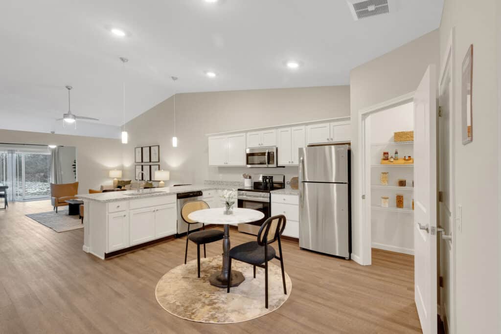 Modern kitchen with white cabinets, stainless steel appliances, a round table with two chairs, and an open pantry displaying various dry goods. Living area visible in the background.