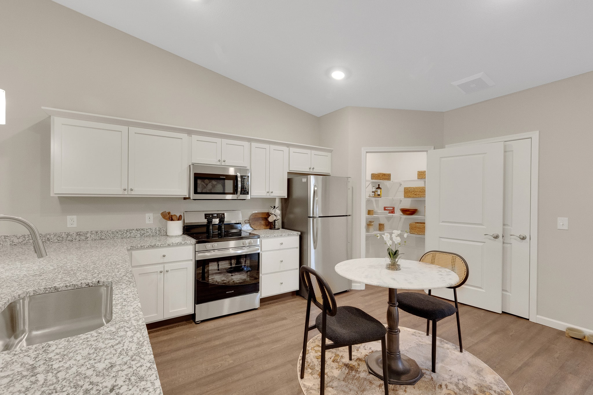 Modern kitchen with white cabinets, stainless steel appliances, and a small round dining table with two chairs on a rug. Open door reveals pantry shelves with baskets.
