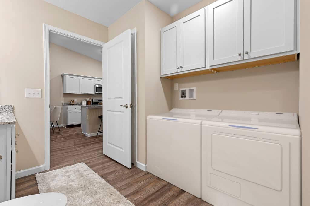 A laundry room with a washer and dryer, white cabinets above, beige walls, and a view into a kitchen with similar cabinets and wood flooring.