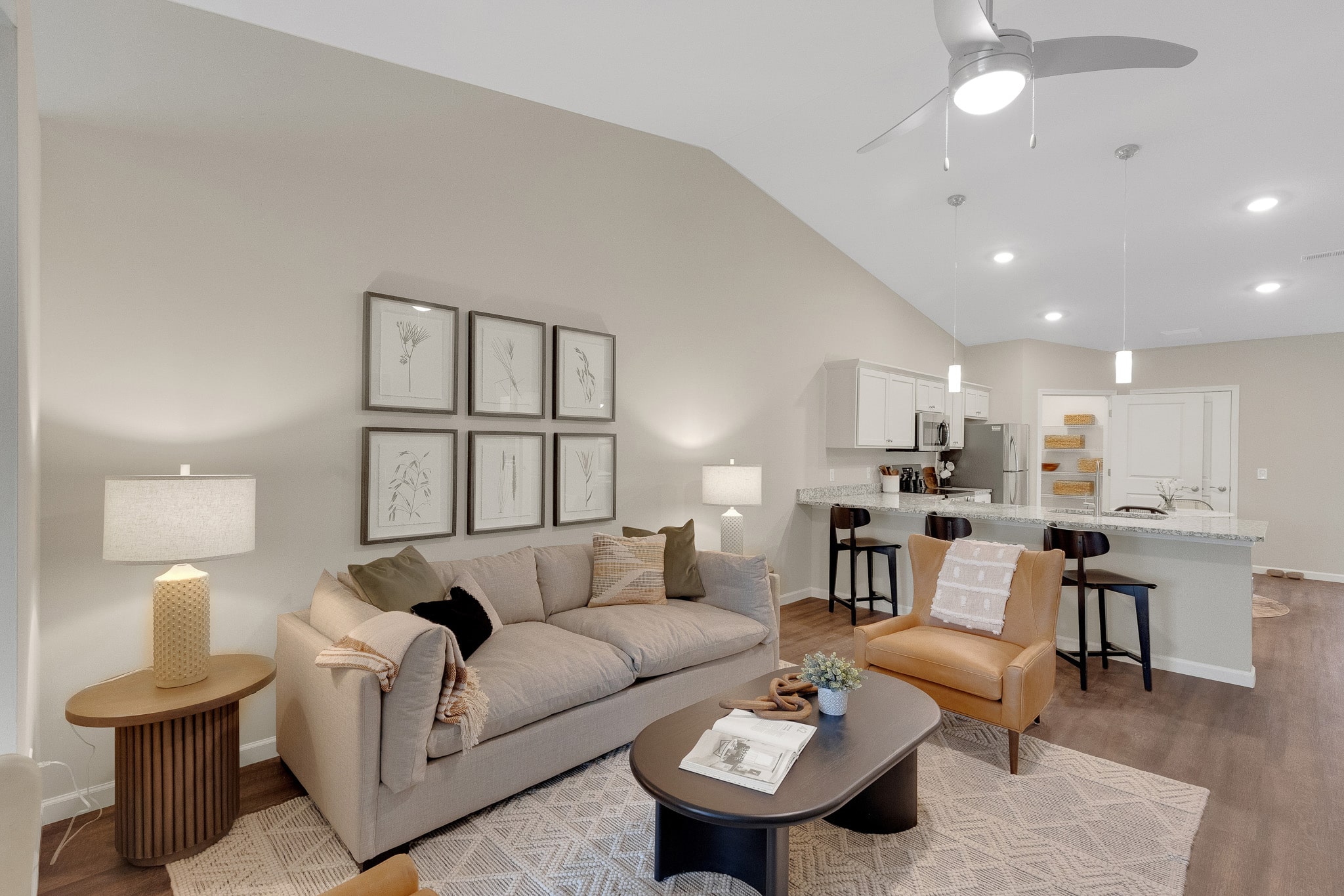 Neutral-toned living room with a beige sofa, coffee table, wall art, two chairs, and a kitchen area in the background under pendant lights.