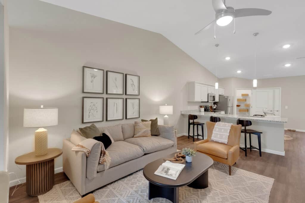 Modern open-concept living room and kitchen with neutral tones, a beige sofa, armchairs, framed botanical art, and a ceiling fan.