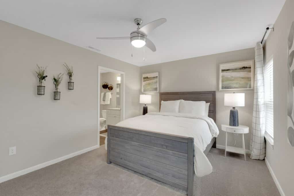 A bedroom with a wooden bed, white bedding, two nightstands with lamps, wall art, and an open door leading to a bathroom. Neutral-colored walls and carpeted floor.