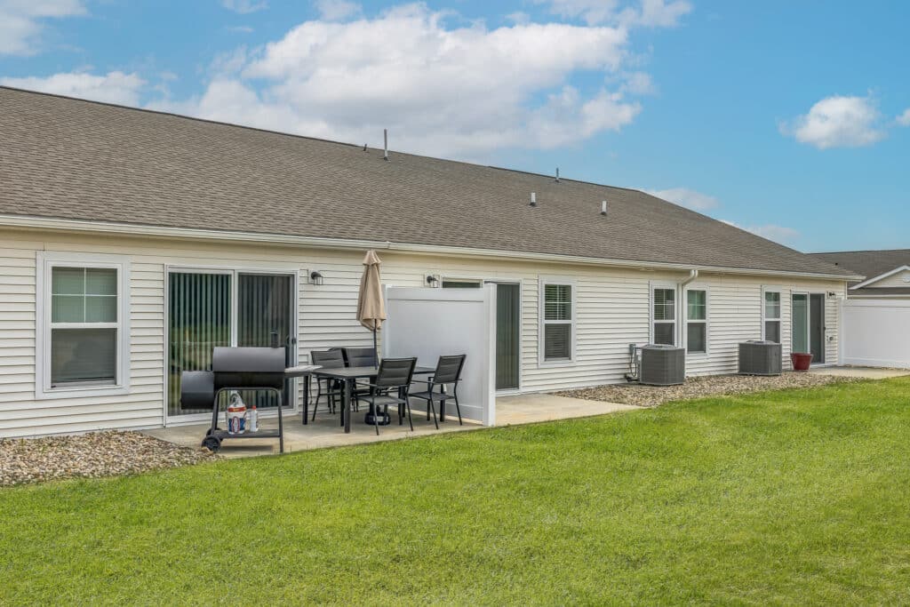Single-story building with white siding, sliding glass doors, patio area with a grill, table, and chairs, next to an air conditioning unit and green lawn under a partly cloudy sky.