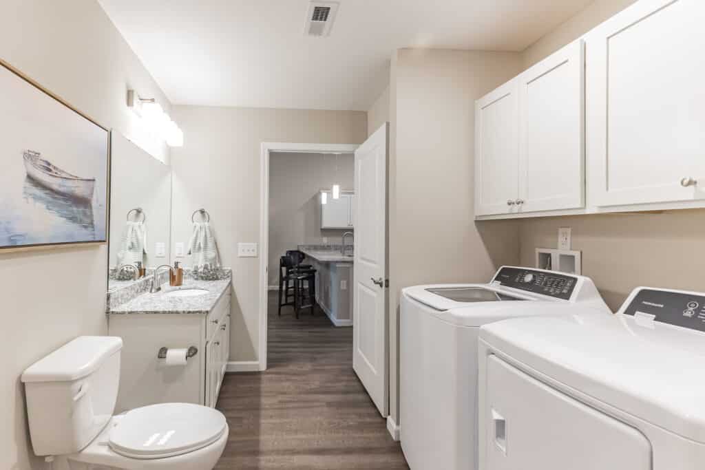 A bathroom with a toilet, double sink vanity, washer, and dryer; white cabinets, granite countertops, and wood flooring, connected to another room through an open doorway.