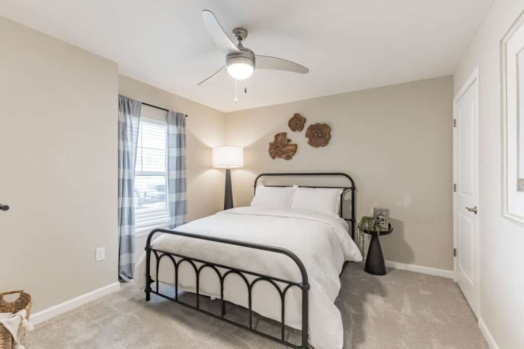 Bedroom with a metal bed frame, white bedding, bedside tables, a floor lamp, wall art, carpeted floor, and a window with checkered curtains.