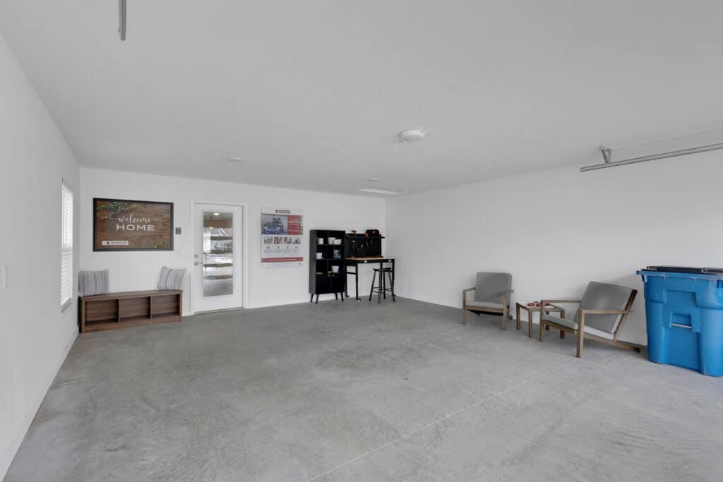 Spacious garage with concrete floor, white walls, two gray chairs, a blue recycling bin, wall posters, a shelving unit, and a bench near a door and window.