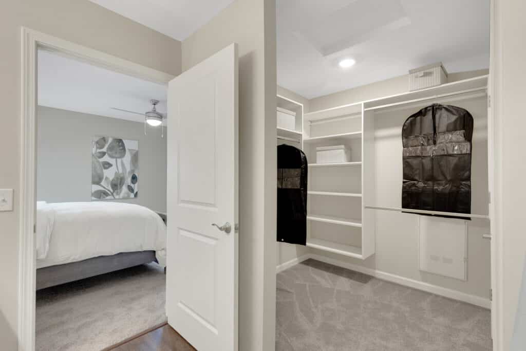 Open walk-in closet with built-in shelves and hanging storage, adjacent to a bedroom with neutral decor and a ceiling fan.