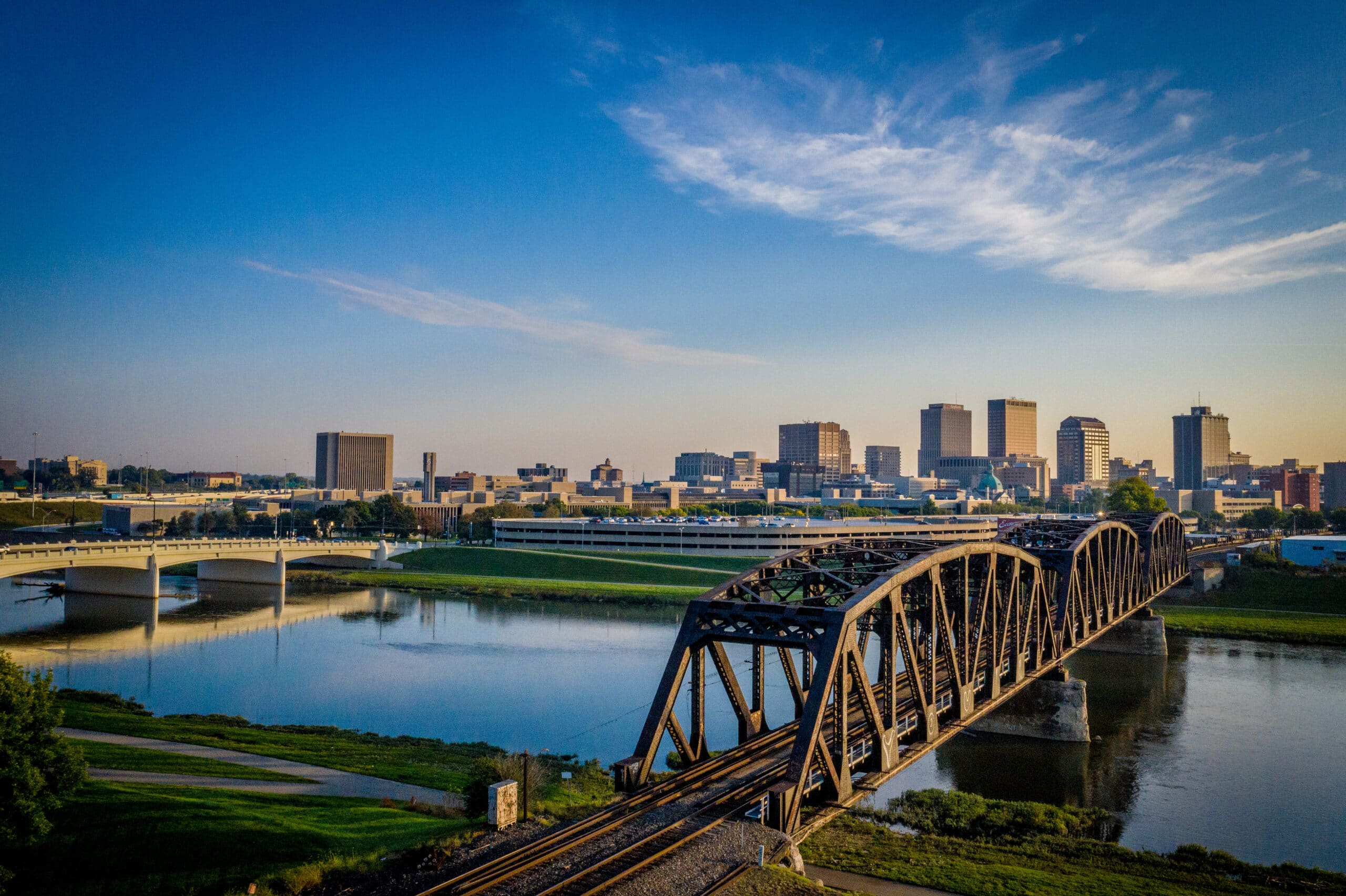 A steel railroad bridge crosses a river toward a city skyline in the background, with modern buildings under a clear blue sky.