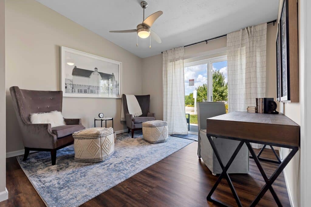 Bright sitting room with two gray armchairs, round poufs, a rug, desk, and large window with white curtains overlooking greenery.