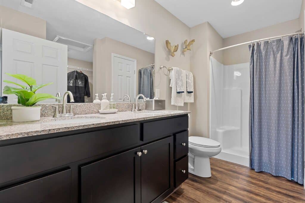 Modern bathroom with a double-sink vanity, granite countertop, potted plant, folded towels, toilet, and a shower with a blue curtain on a wood-look floor.