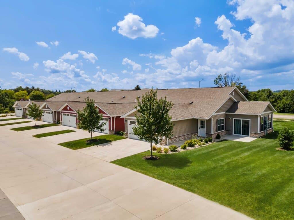 Single-story townhomes with attached garages and well-maintained lawns on a sunny day, surrounded by trees and a clear blue sky with scattered clouds.