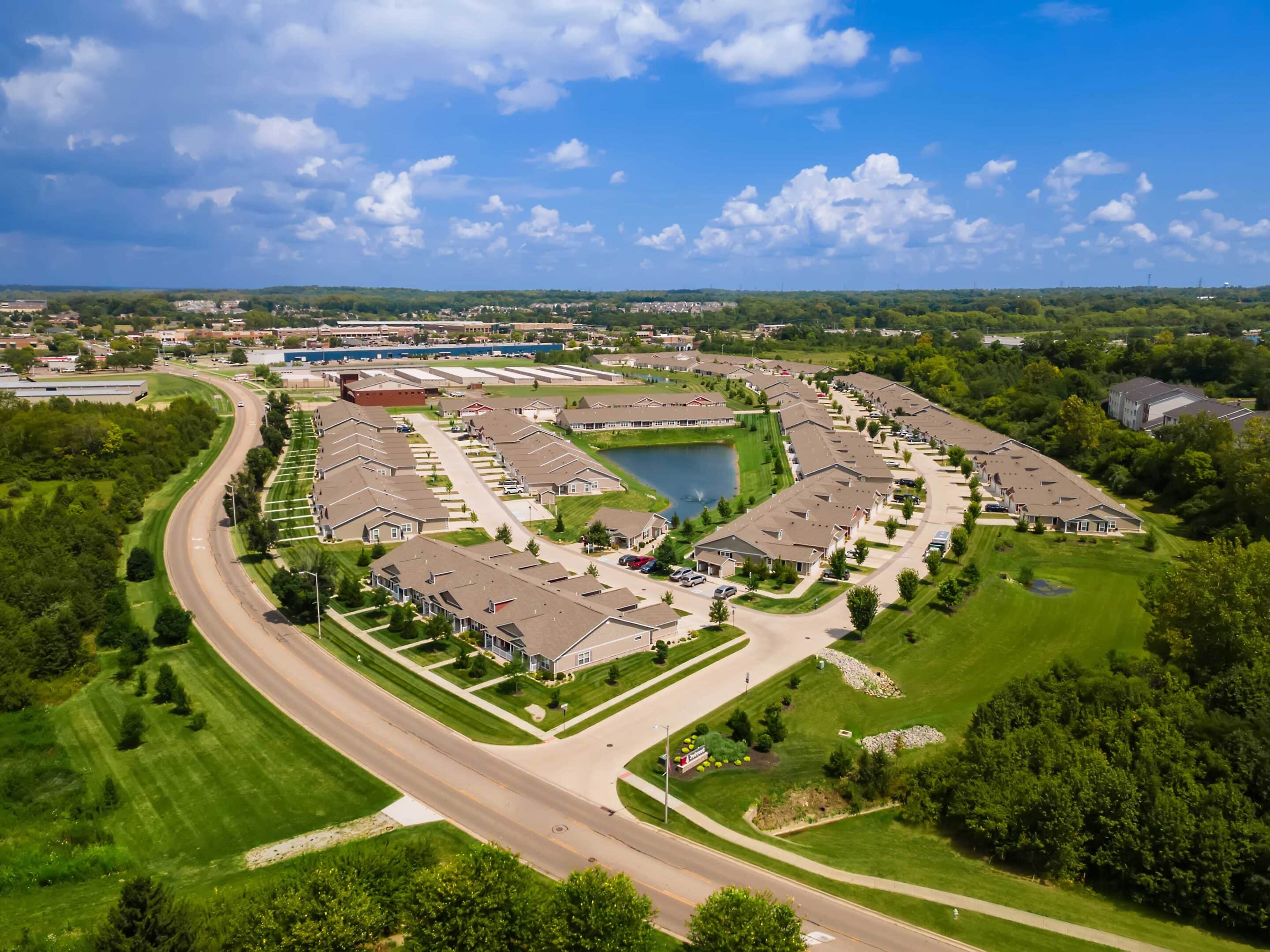 Aerial view of a suburban neighborhood with rows of houses, curved streets, green lawns, and a central pond under a blue sky with scattered clouds.