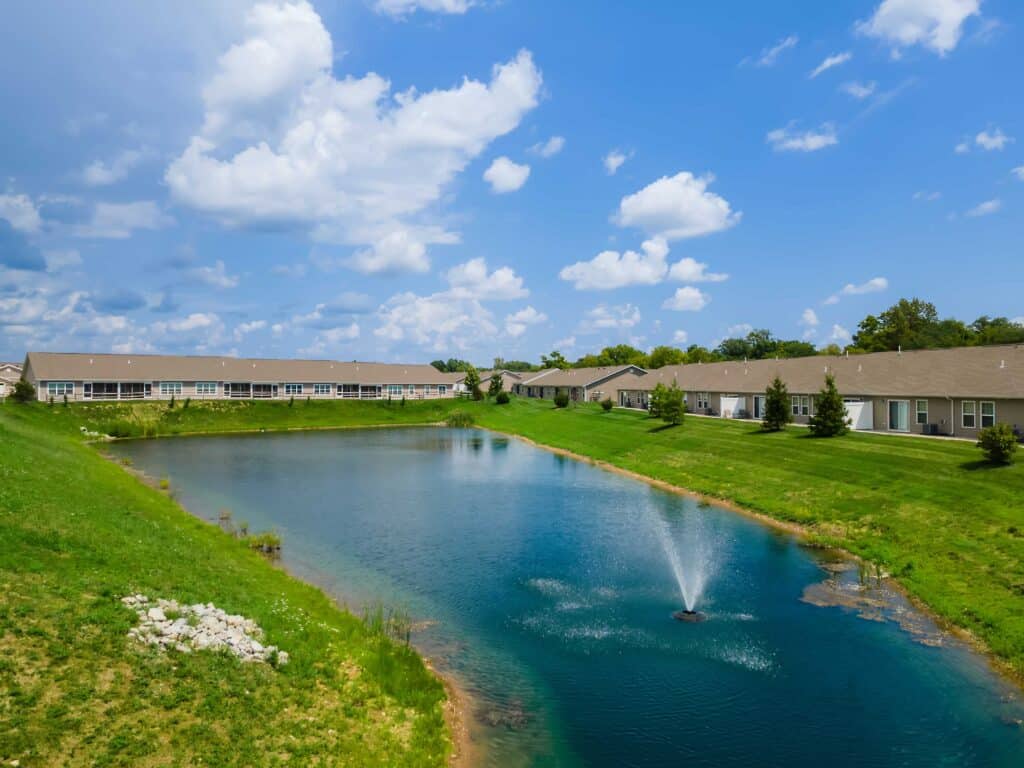 A small pond with a fountain is surrounded by grassy banks and single-story residential buildings under a partly cloudy sky.