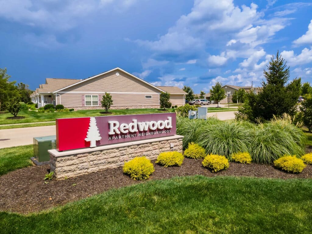 A red and white sign reading "Redwood Apartment Neighborhoods" stands in front of landscaped bushes and a residential building under a partly cloudy sky.