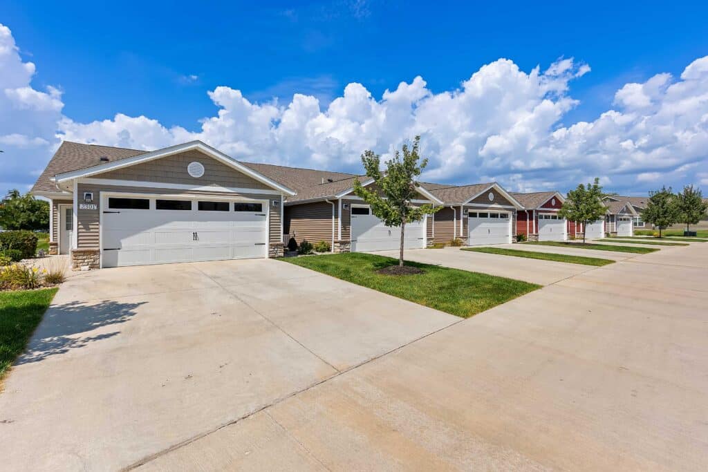 Row of single-story suburban houses with attached garages, driveway, and small front lawns on a clear, sunny day.