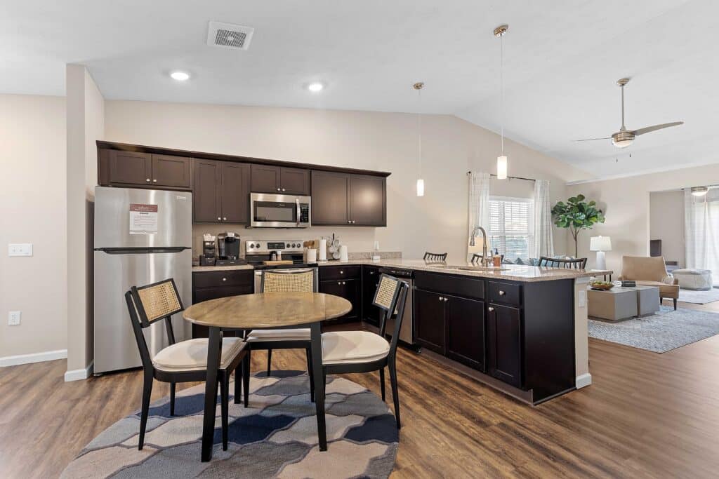 Modern kitchen with dark cabinets, stainless steel appliances, a round dining table with four chairs, and an open layout leading to a living area with a ceiling fan and large windows.