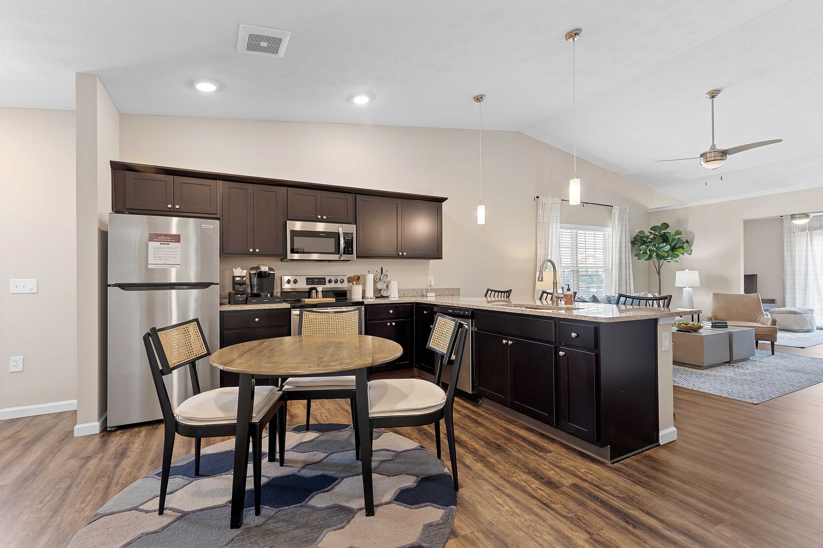 Modern kitchen with dark cabinets, stainless steel appliances, a round dining table with four chairs, and an open layout leading to a living area with a ceiling fan and large windows.