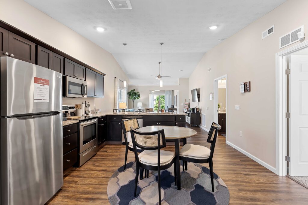 Modern kitchen and dining area with dark cabinets, stainless steel appliances, round table with four chairs, and wood-style flooring in an open-plan layout.